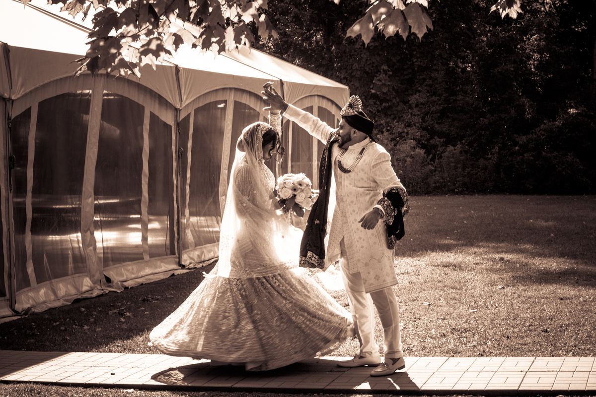 A bride and groom dance joyfully outdoors, the bride's lehenga blurring as she twirls near a wedding tent.