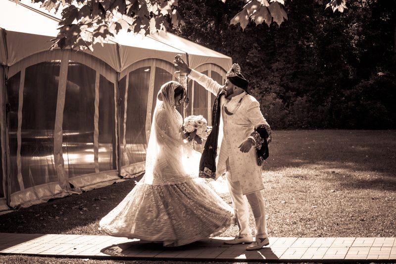 A bride and groom dance joyfully outdoors, the bride's lehenga blurring as she twirls near a wedding tent.