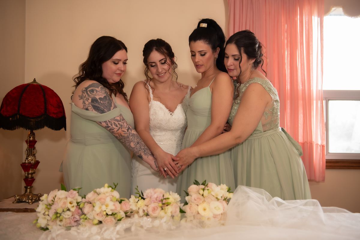 Four women, including a bride in white, gather around a table with flowers and a vintage lamp, hands clasped, in warm light.