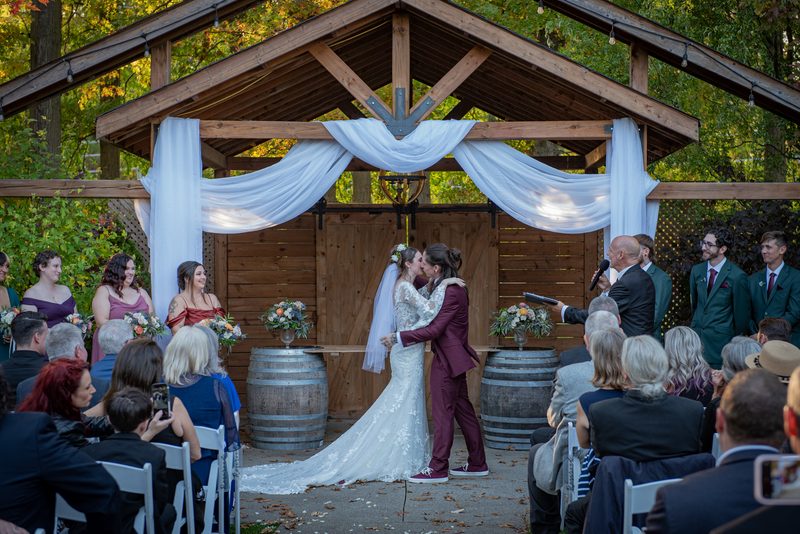 Bride and groom kiss beneath wooden arbor draped with white fabric, flanked by wine barrels and flowers.