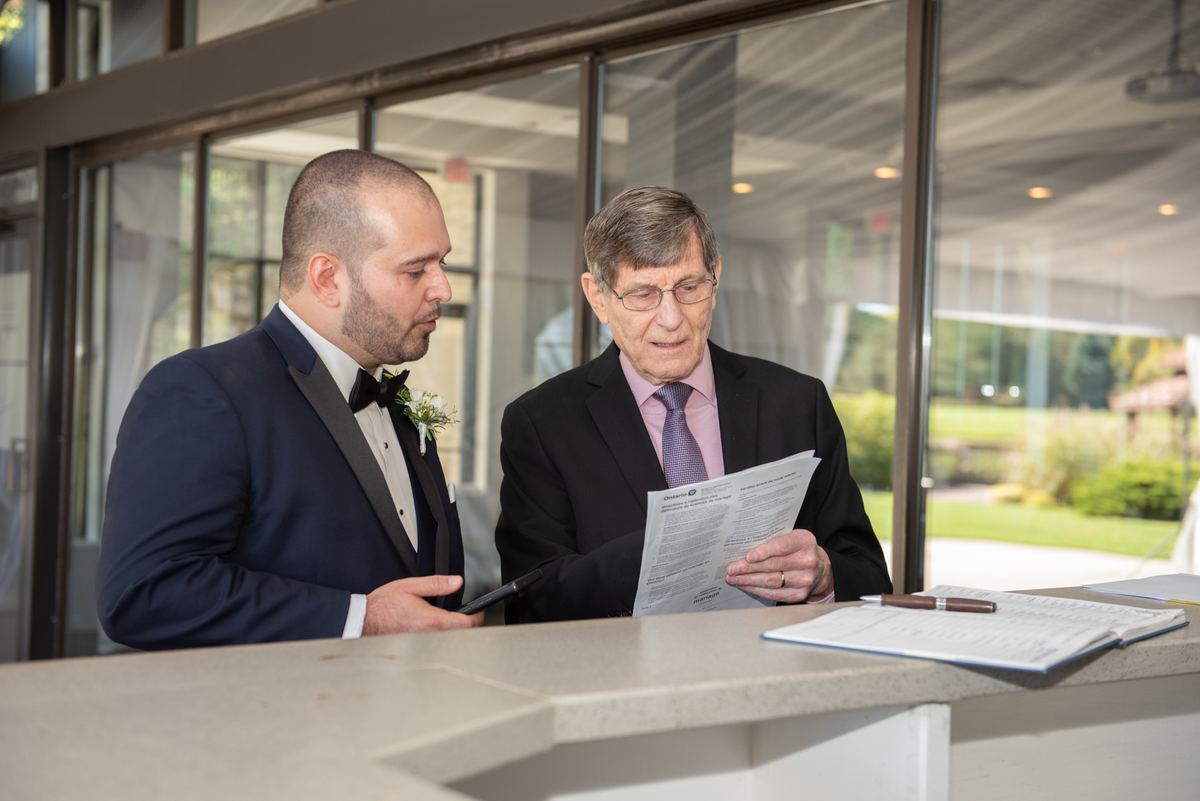 Two formally dressed men review documents at a desk illuminated by natural light from windows behind them, papers.