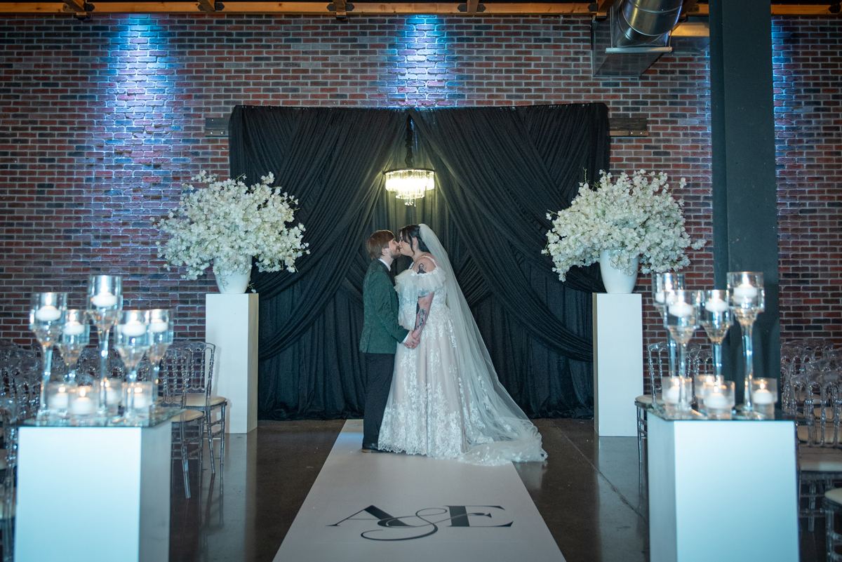 Bride and groom share a kiss at an altar with blue spotlights illuminating brick wall and black drapery.