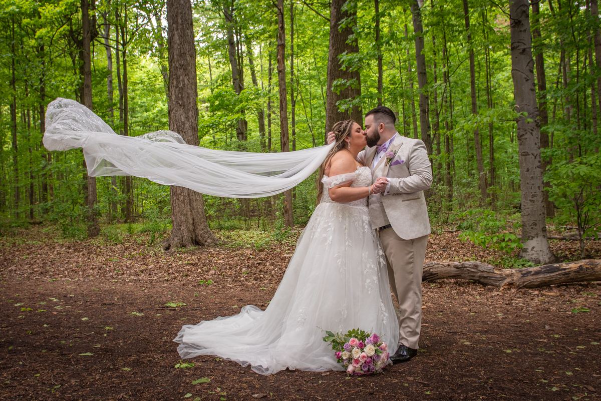 A bride and groom kiss in a sunlit forest, their hands intertwined, with her veil fluttering in the breeze.