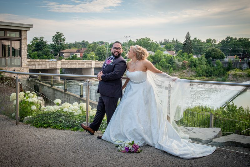 A smiling bride in a white gown and veil stands with her groom in a dark suit by a railing overlooking a river.
