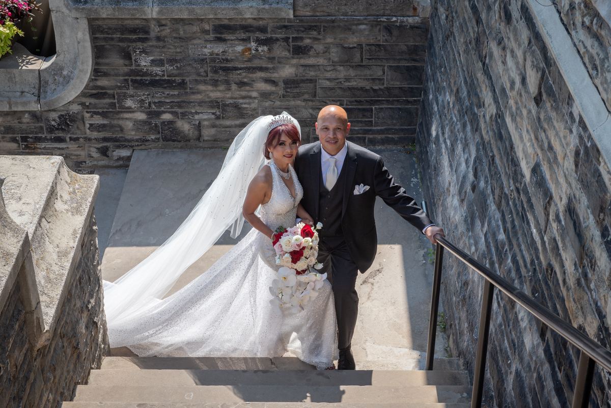A newlywed couple in bright sunlight, the bride in a white gown and red and white flowers, the groom in a dark suit.