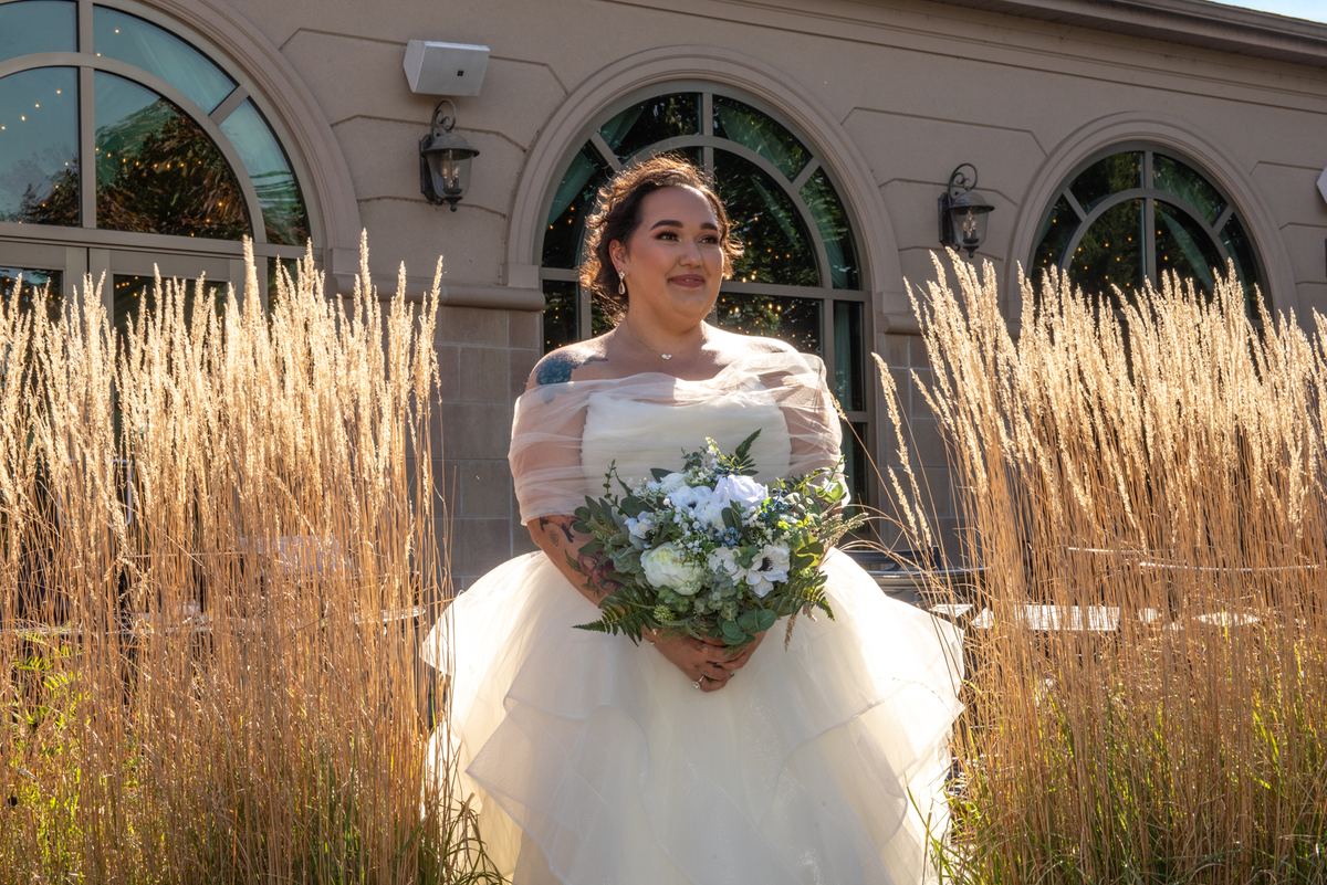 A bride in an elegant off-the-shoulder gown stands in a field of golden grass, holding white and blue flowers.