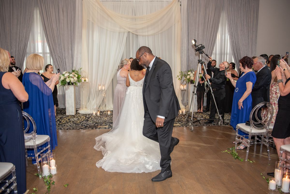 The bride and groom embrace warmly as they walk down the aisle surrounded by guests taking photos, with elegant drapery.