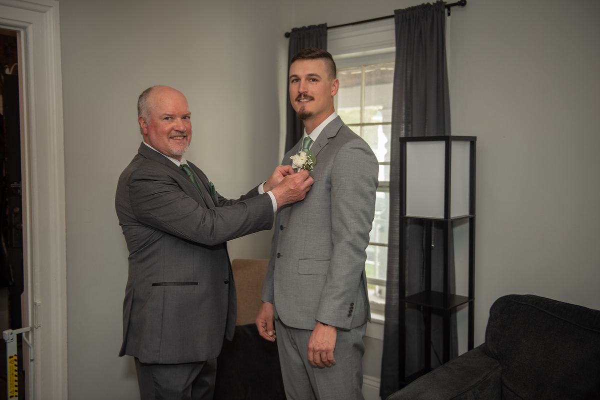 An older man pins a white boutonnière onto the lapel of a younger man in a gray suit, smiling warmly at each other.