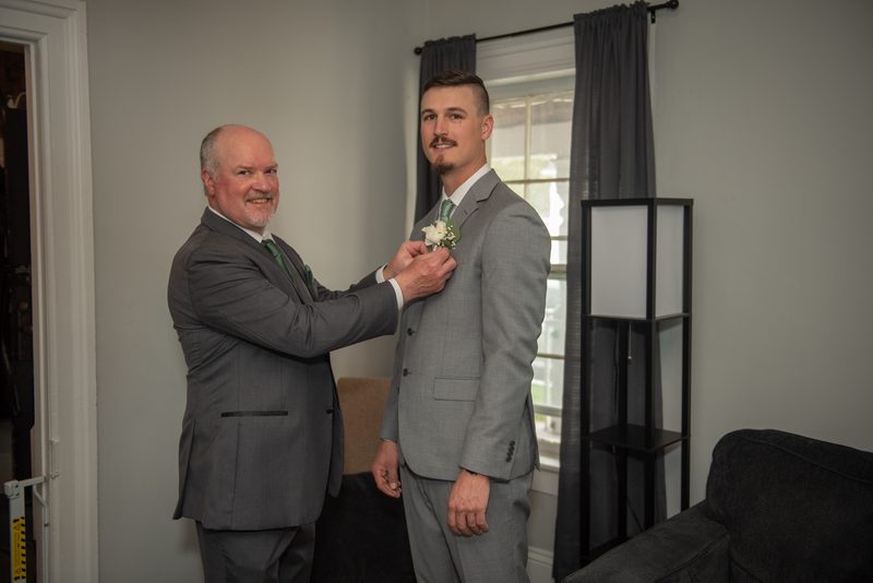 An older man pins a white boutonnière onto the lapel of a younger man in a gray suit, smiling warmly at each other.