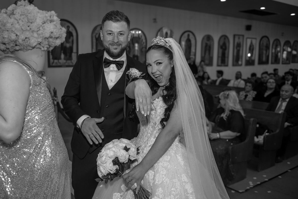Bride laughing exuberantly as groom smiles warmly, holding her bouquet and his arm around her in church.