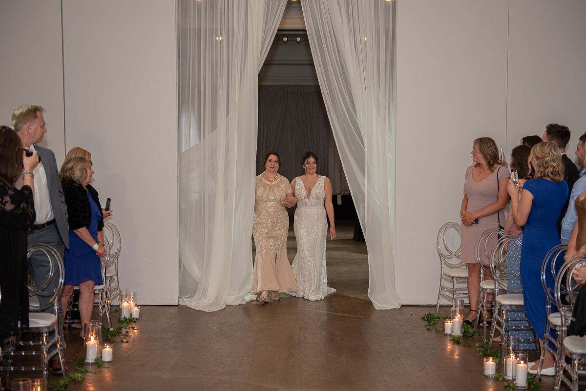 Two brides walk hand-in-hand down a candlelit aisle toward the ceremony space, framed by sheer white curtains.