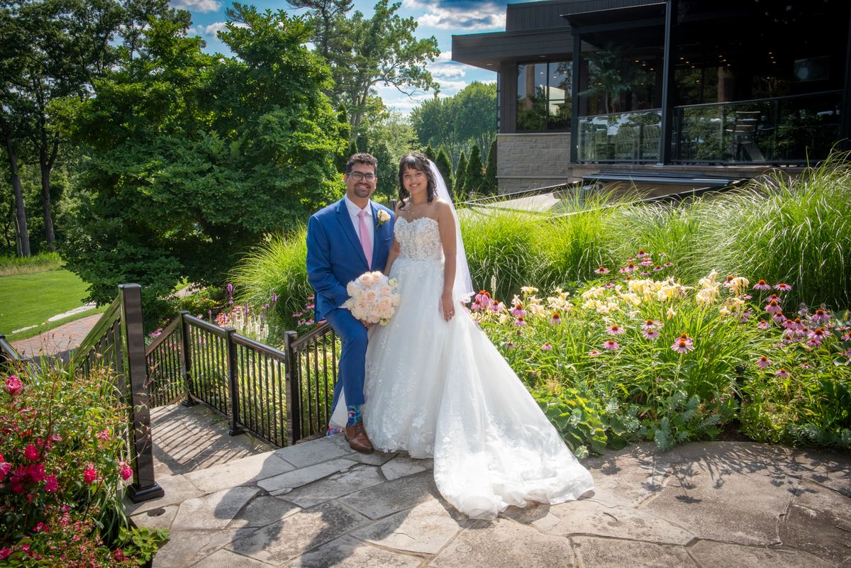 A smiling bride and groom pose on a stone pathway surrounded by gardens, the groom sitting on a railing.