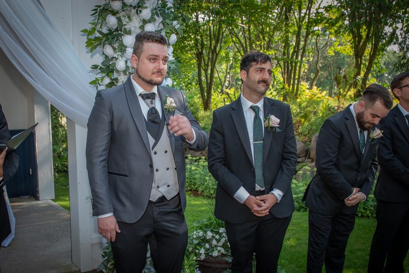 Groomsmen in formal suits stand solemnly under a floral arch during an outdoor wedding ceremony, their emotions evident.