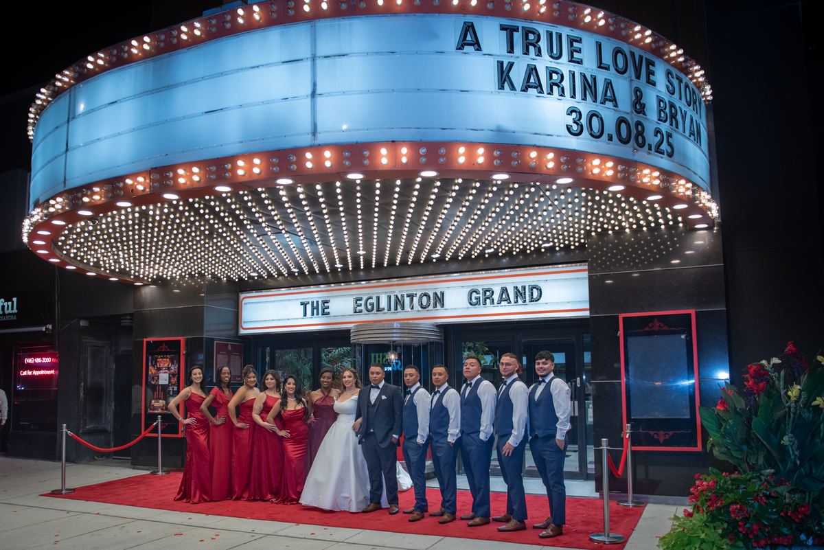 A bride and groom stand with their wedding party in front of an illuminated marquee at a theater, under vintage-style lights.