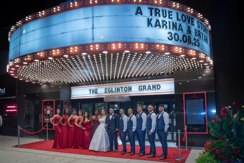 A bride and groom stand with their wedding party in front of an illuminated marquee at a theater, under vintage-style lights.