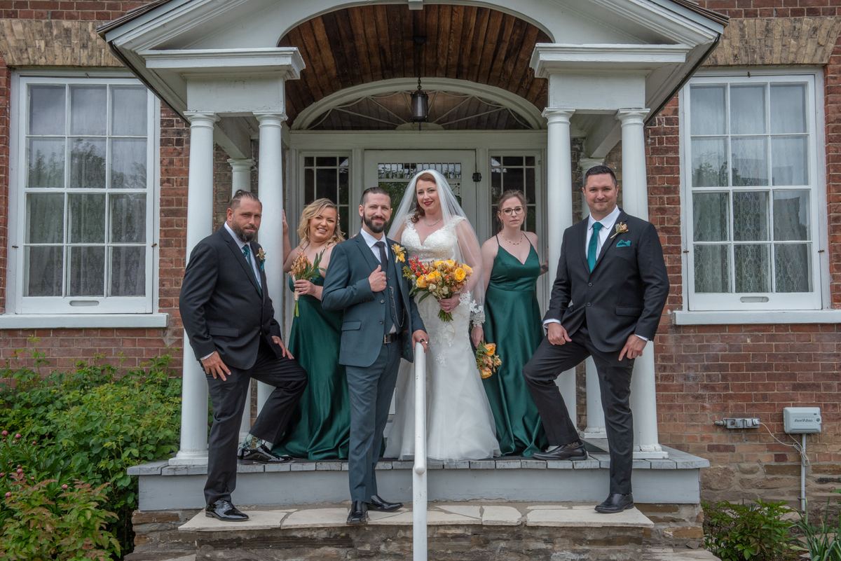 A wedding party poses on stone steps with a white-columned porch behind, holding bouquets of yellow and orange flowers.