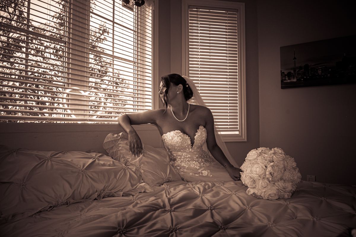 A bride in her wedding gown sits pensively by a window with striped light and her bouquet beside her.