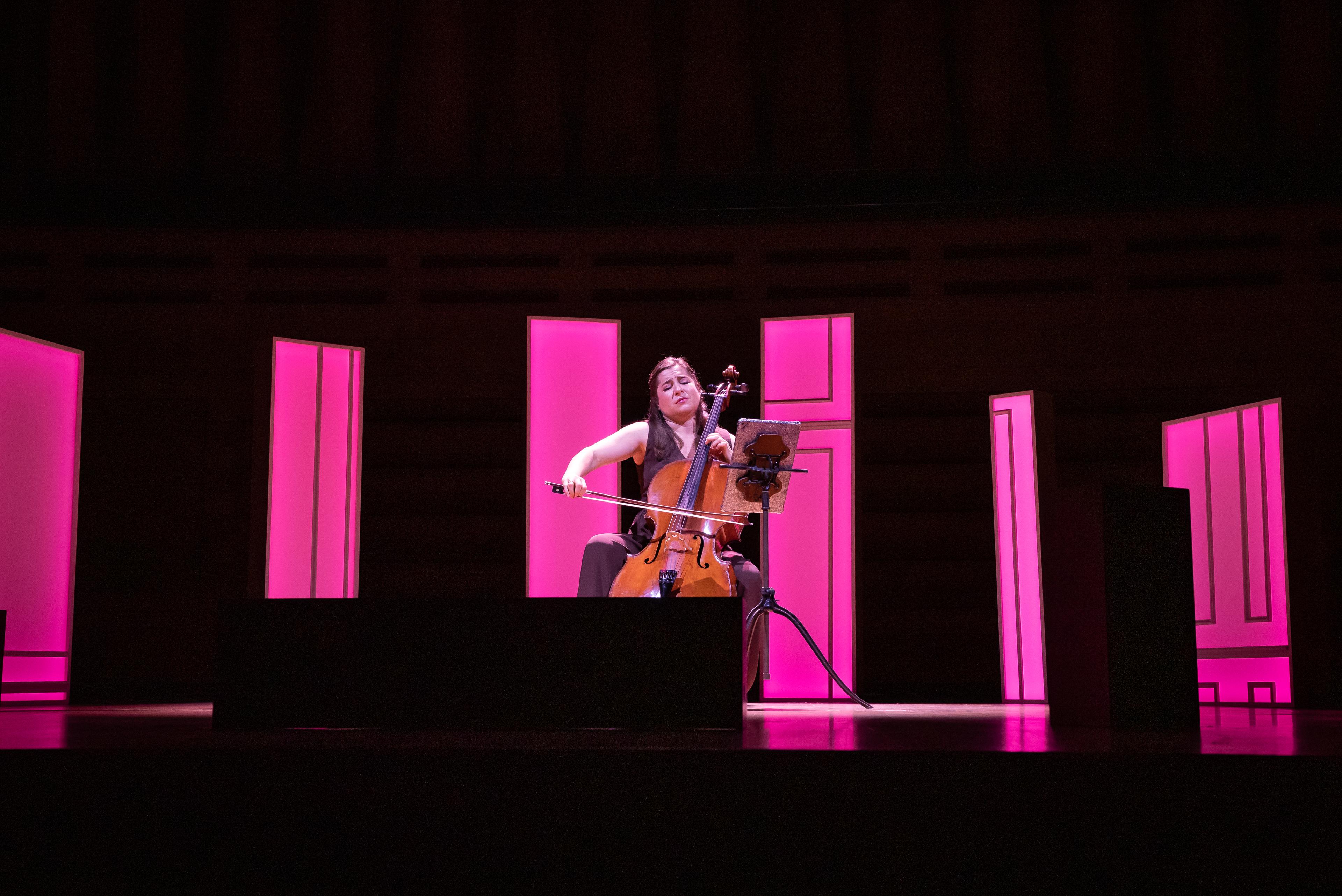 Cellist Alisa Weilerstein playing solo in the middle of a circle of wooden set pieces, all different sizes, illuminated pink.