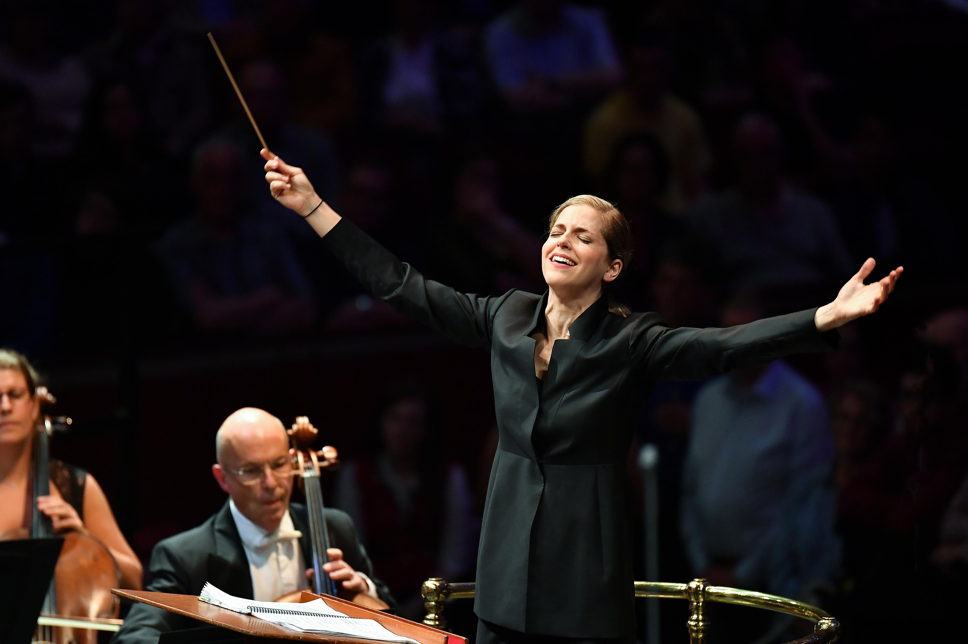 Karina Canellakis, a white woman with blonde hair wearing a black suit, in front of a orchestra with her arms out stretched
