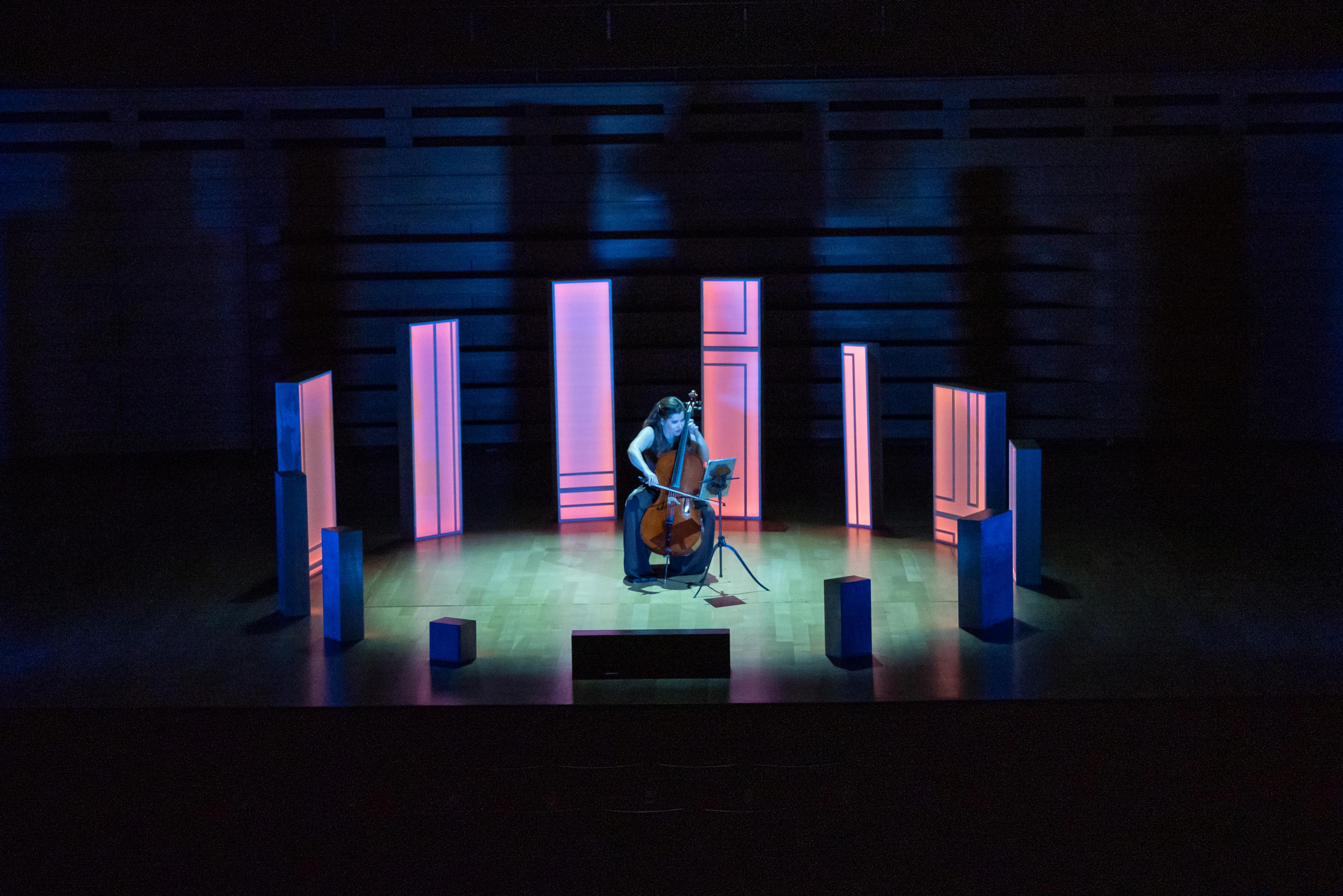 Cellist Alisa Weilerstein playing solo in the middle of a circle of wooden set pieces, all different sizes, illuminated pink.