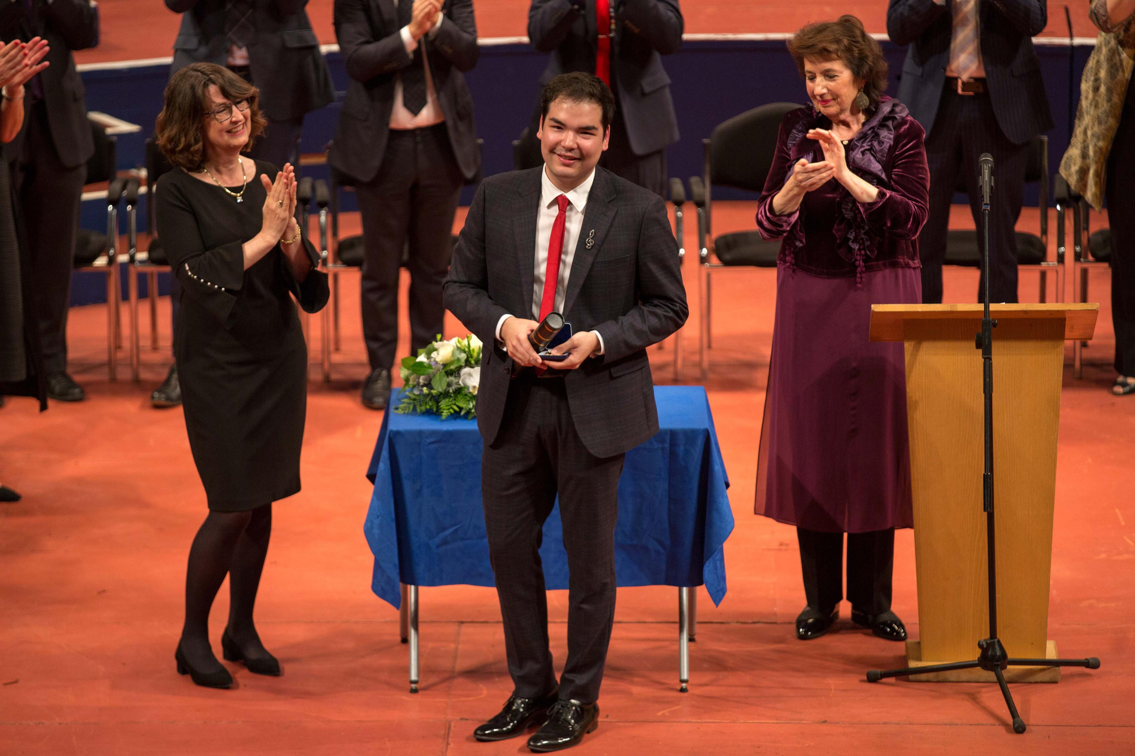 Alim Beisembayev stood wearing a suit on stage receiving his gold medal from the 2021 event, receiving applause from 2 ladies stood either side of him