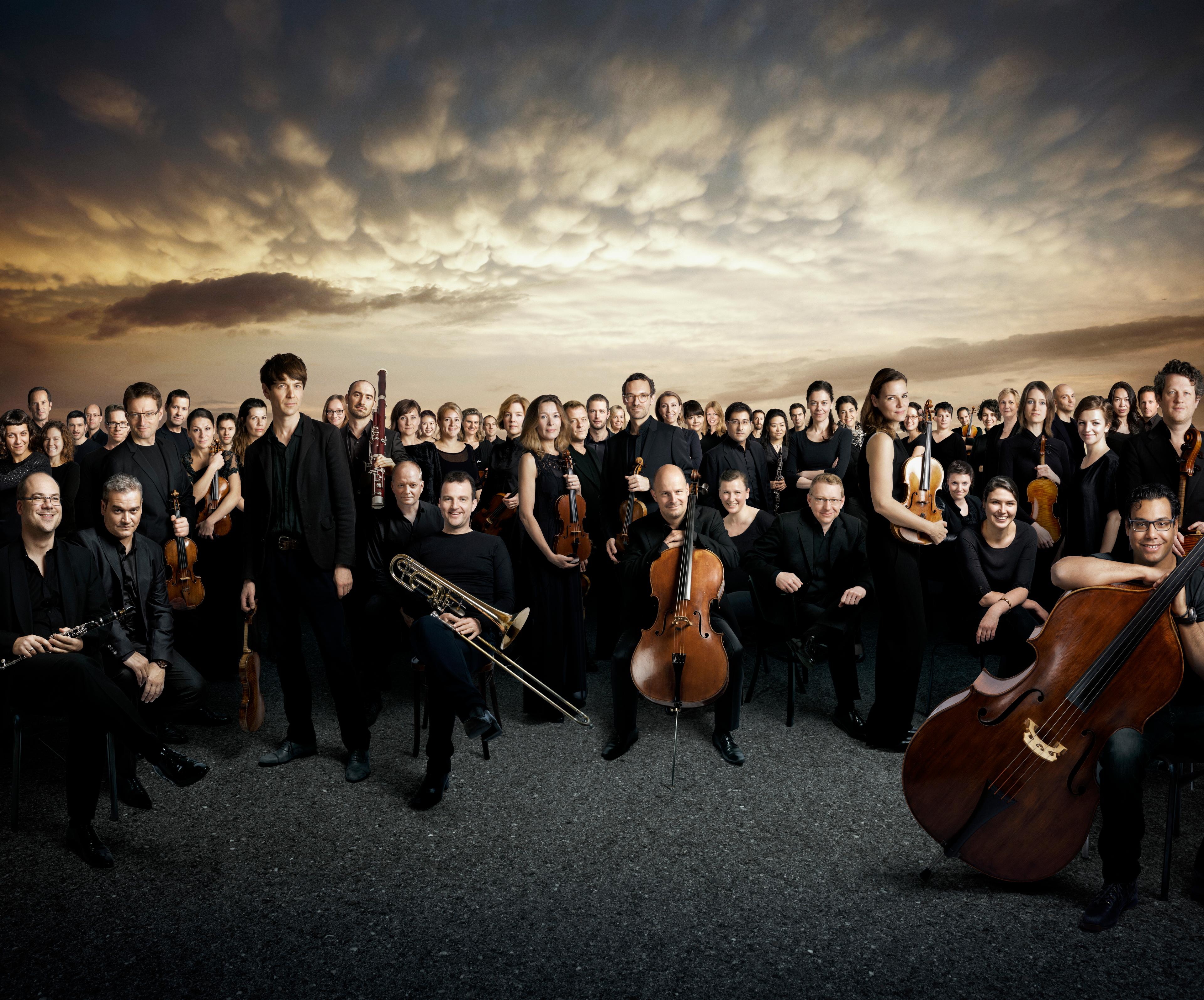 The orchestra wearing all black posed looking at the camera against a dramatic sky 