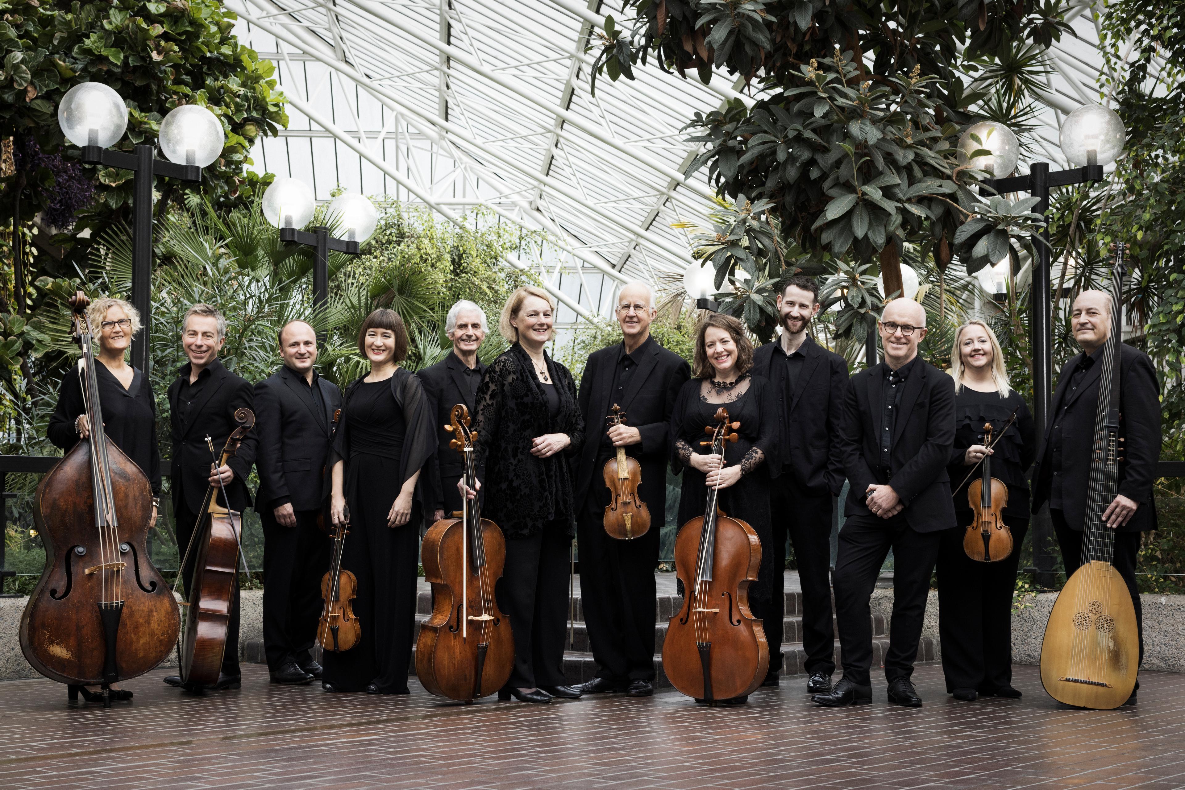 The Academy of Ancient Music standing with their instruments in a greenhouse smiling at the camera with foliage behind them