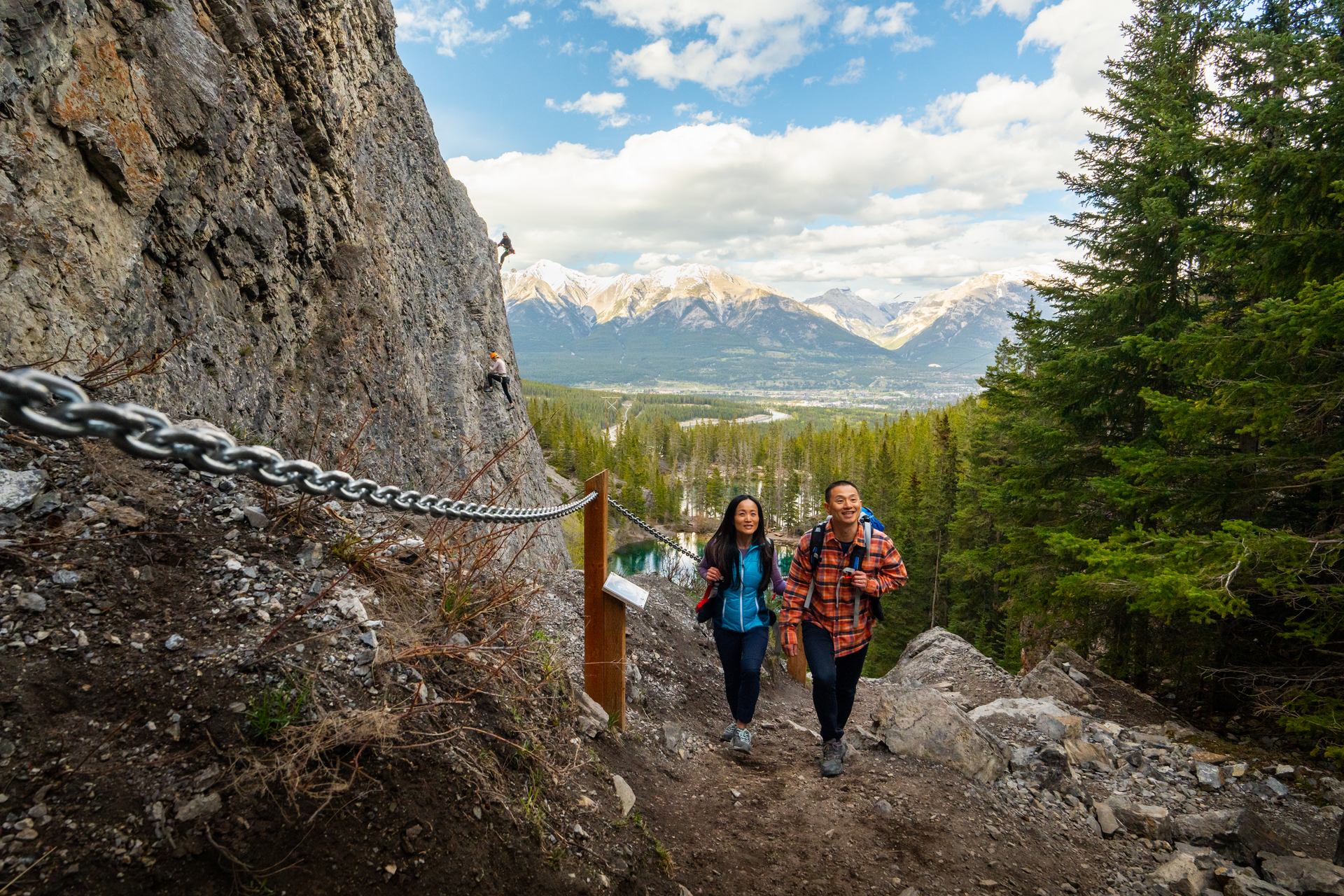 A couple walking up the Grassi Lakes Trail with rock climbers in the distance.