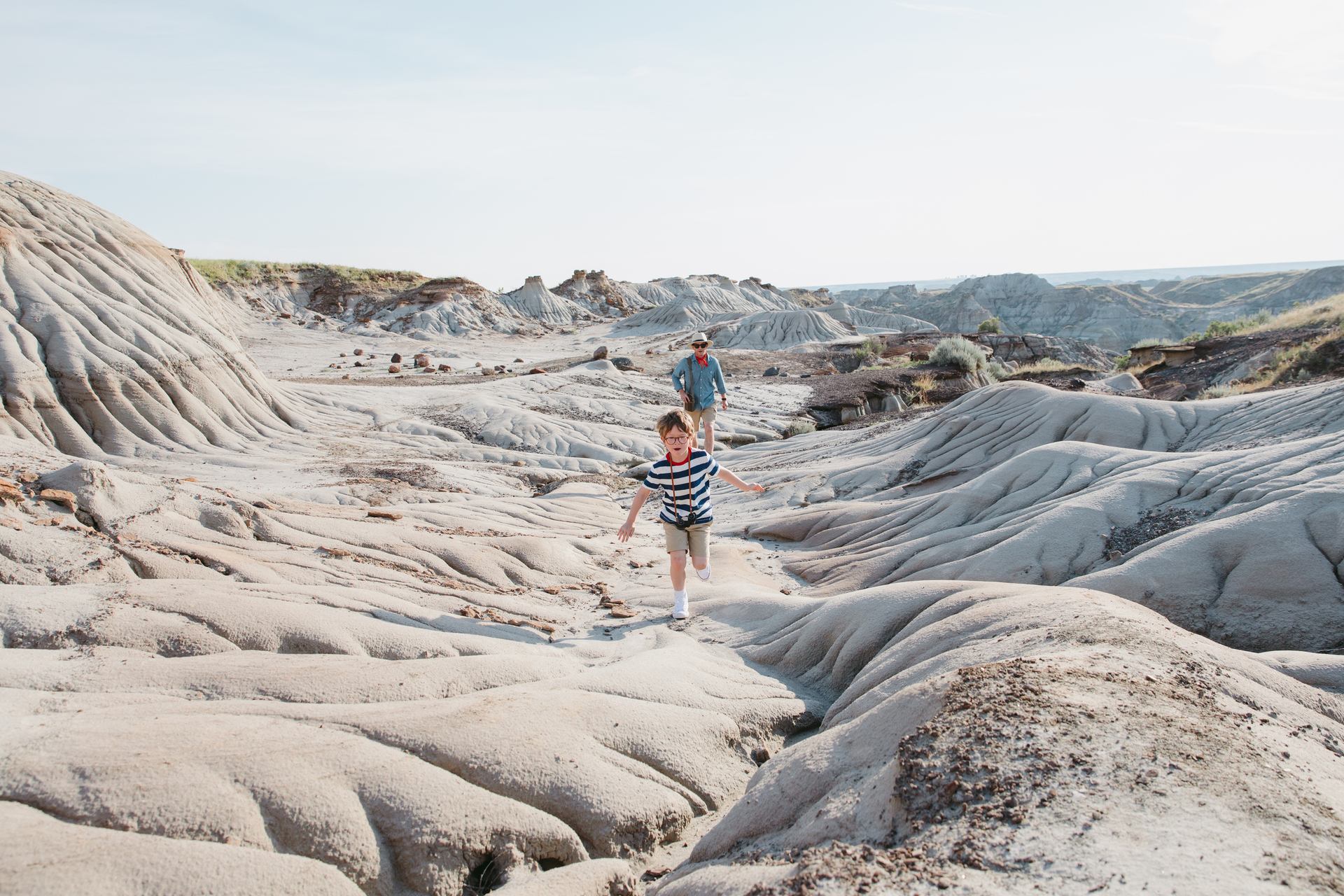 A little boy runs along rock coulees in the Canadian badlands, with his parent standing behind him.