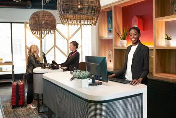 Receptionist smiling behind a modern reception desk while her colleagues checks in a guest with luggage beside her.