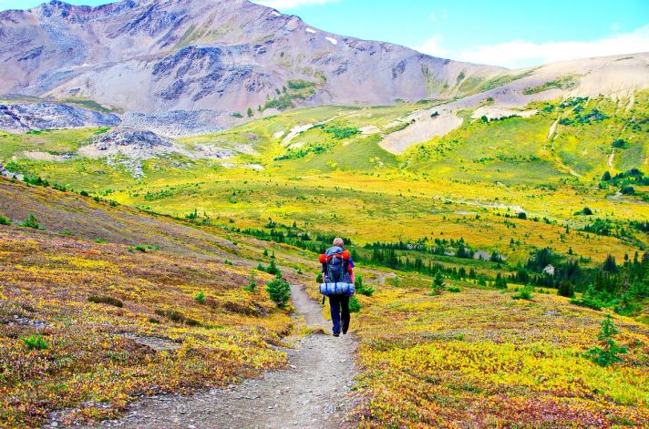 Backpacker with gear hiking on a trail toward the mountains in the background.