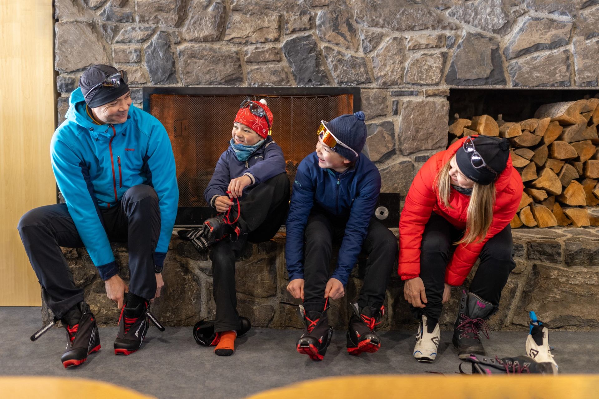 A family of four putting on their ski boots while sitting on a ledge in front of a fireplace.