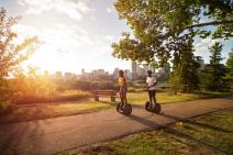 Two people ride Segways down a path as the sun goes down, Edmonton skyline in the background