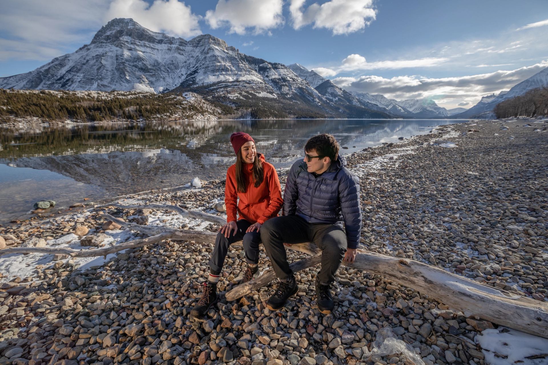 A couple take a rest on a fallen tree with a lake and mountains in the background.