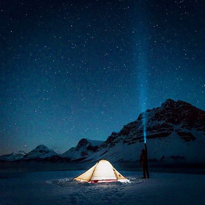 A person beside a tent at night, shining a headlamp into the dark starry sky while winter camping in the mountains.