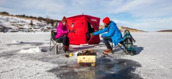 A couple sitting in front of a portable ice hut, with a fire going while ice fishing.