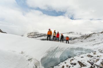Group of four smiling as they follow a tour guide along a glacier walk in the snow covered mountains.