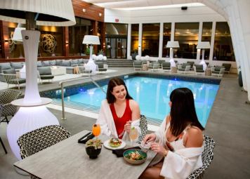 Two women in bathing attire sitting poolside enjoying drinks and food, making for a very trendy atmosphere