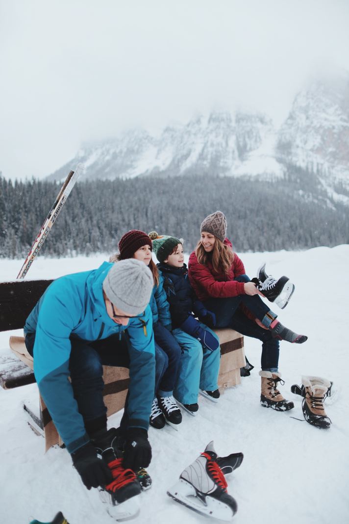 A family of four sitting on a bench to lace up their ice skates with the frozen lake and mountain view in the background.