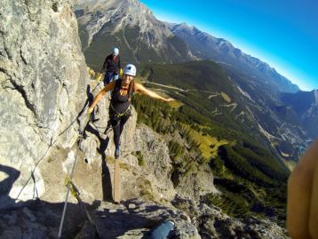 Two climbers smiling as they walk across a narrow plank while holding rope along the side of a mountain with a valley view in the background.