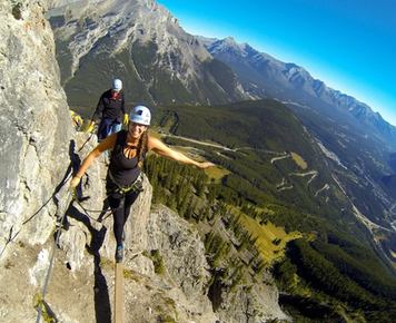 Two climbers smiling as they walk across a narrow plank while holding rope along the side of a mountain with a valley view in the background.
