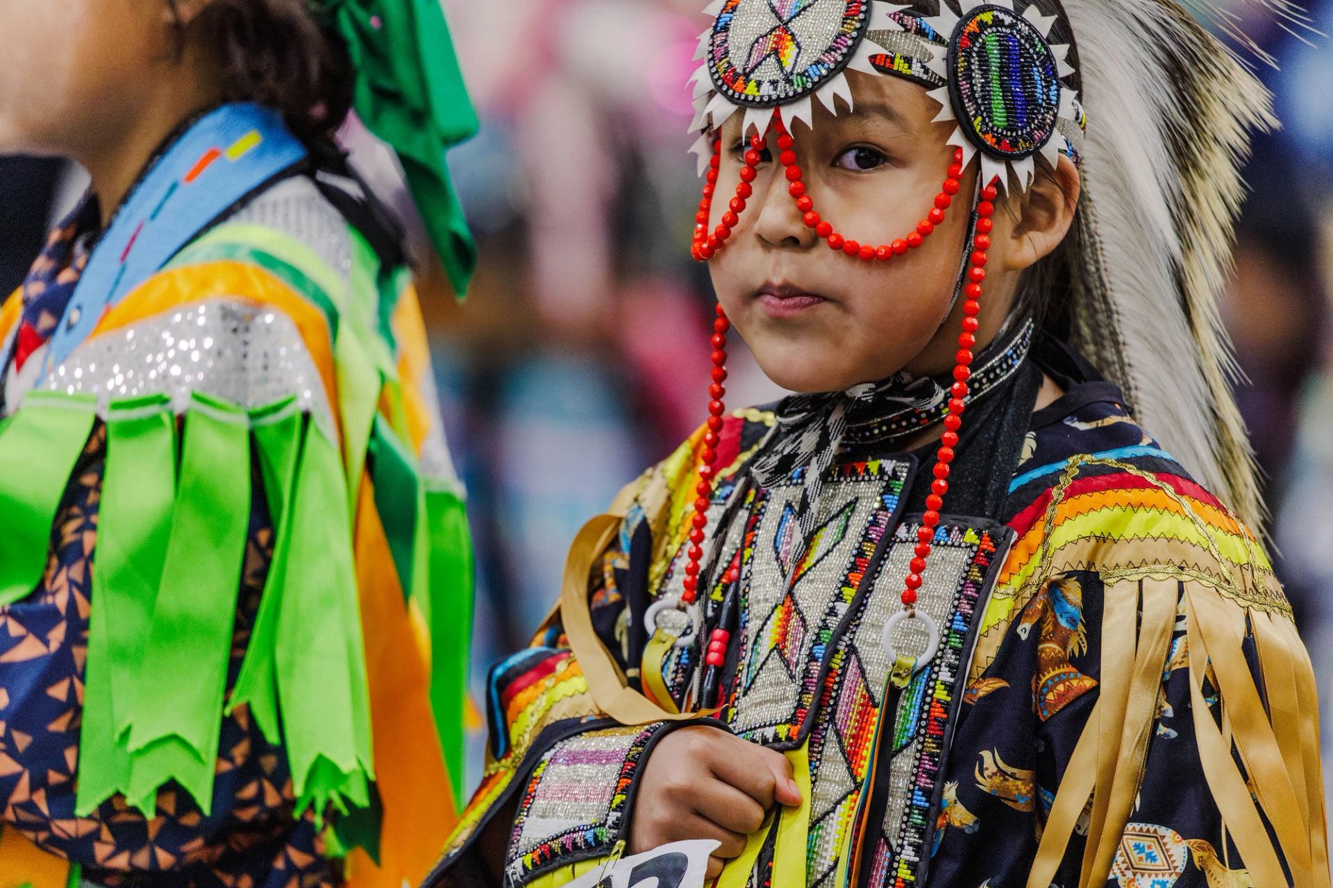 A child in colourful regalia, including an embroidered headband, stands calmly during a powwow.