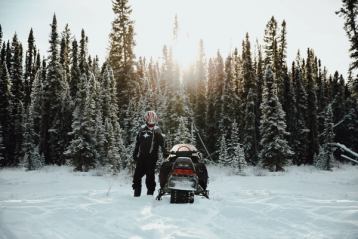 A man standing beside his snowmobile with the soft sun coming through the trees while snowmobiling.