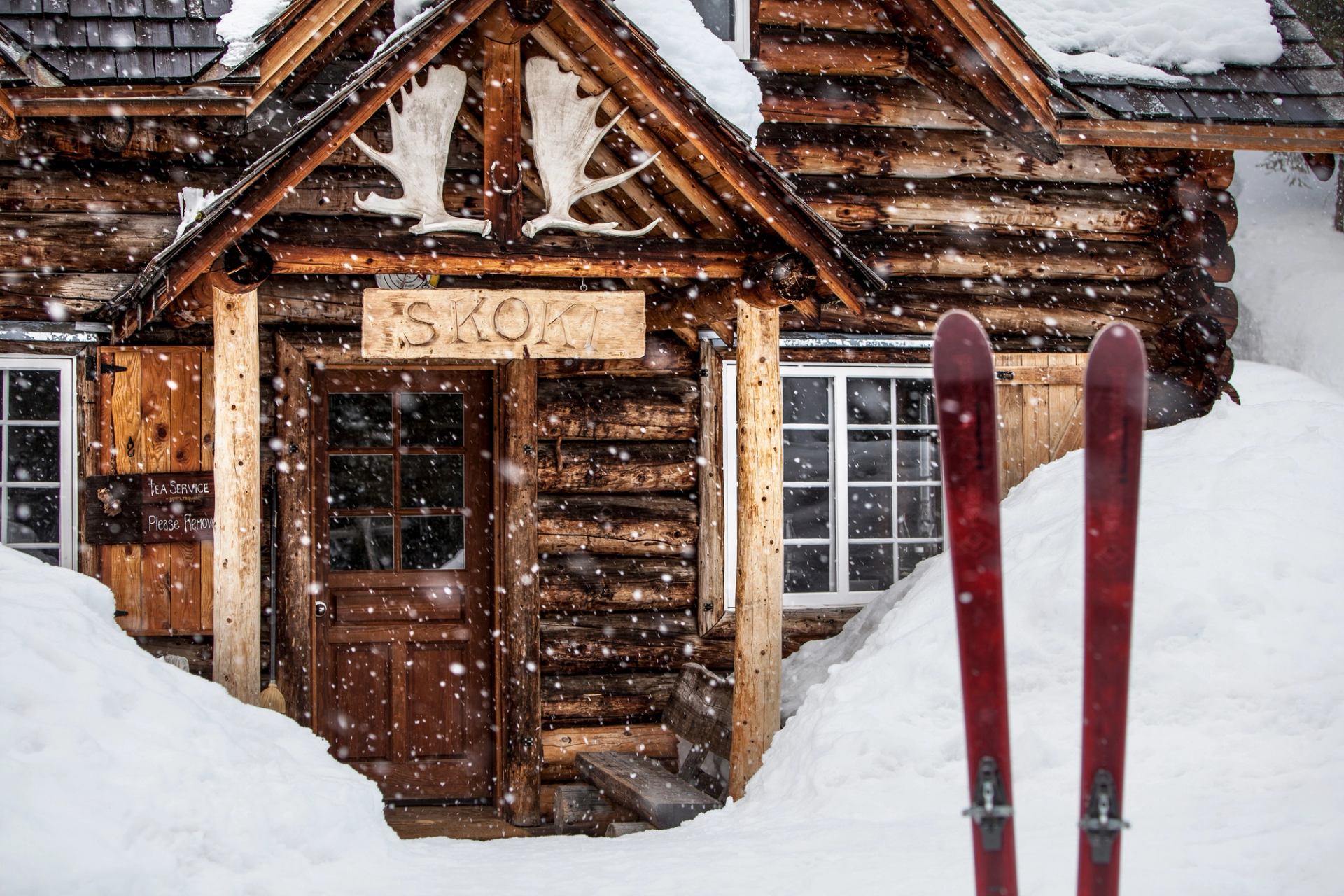 The exterior of Skoki Lodge, a log cabin, is surrounded by tall snow drifts and fluffy falling snowflakes.