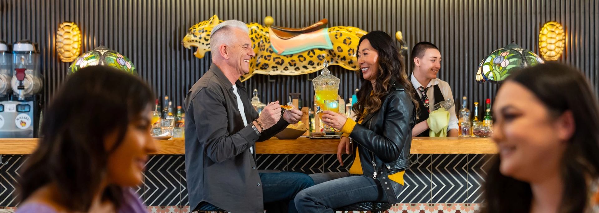 A woman holds a drink and smiles at a man she is sitting with at the bar as he prepares food to eat. In the foreground; two woman are having a conversation over food and drinks.