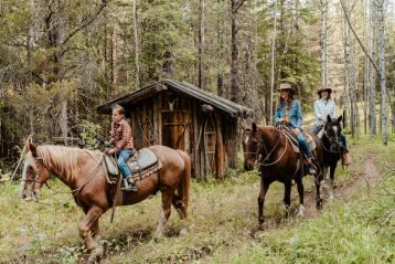 People ride horses past a wooden shed in the forest.