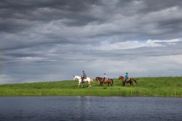 Guests horseback riding beside a pond, dark clouds in the sky, at Rocking R Guest Ranch