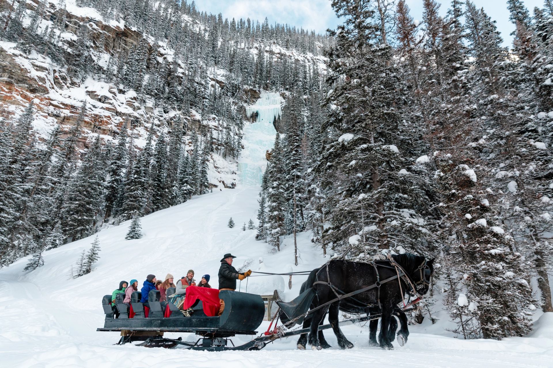 A group of travellers enjoy a horse-drawn sleigh ride along the shore of Lake Louise with a frozen waterfall and forest in the background.