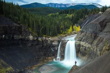 Zoomed out wide shot of a hiker looking at a beautiful waterfall plunging onto sandstone rock below