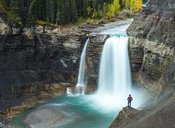 Zoomed out wide shot of a hiker looking at a beautiful waterfall plunging onto sandstone rock below